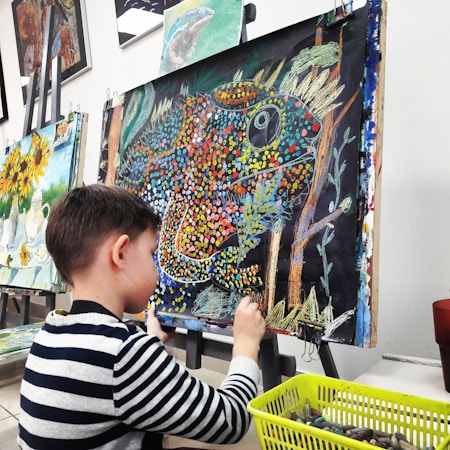 A young child is seated at an easel, working on a colorful painting using oil pastels. The painting features a vibrant depiction of a chameleon with a dotted pattern in various colors such as orange, blue, and yellow, against a dark background. Nearby is a basket filled with art supplies, indicating an active engagement in the creative process. Behind, there are other finished paintings with sunflowers and still life, hanging on a wall.
