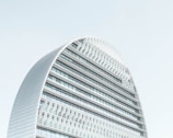 A modern, curved building with multiple horizontal lines and glass panels against a clear sky. The architecture features a sleek, contemporary design with a prominent arch and a grid-like facade.