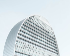 A modern, curved building with multiple horizontal lines and glass panels against a clear sky. The architecture features a sleek, contemporary design with a prominent arch and a grid-like facade.