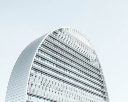 A modern, curved building with multiple horizontal lines and glass panels against a clear sky. The architecture features a sleek, contemporary design with a prominent arch and a grid-like facade.