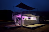 A modern, two-level outdoor structure illuminated with purple lights. The structure has railings on the upper level, a staircase on the side, and a logo with the text 'Wolf Pack Rentals' on the lower level. The surrounding area is dark, suggesting it's night time. There is a flat platform or deck area in front of the structure.