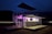A modern, two-level outdoor structure illuminated with purple lights. The structure has railings on the upper level, a staircase on the side, and a logo with the text 'Wolf Pack Rentals' on the lower level. The surrounding area is dark, suggesting it's night time. There is a flat platform or deck area in front of the structure.