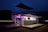 A modern, two-level outdoor structure illuminated with purple lights. The structure has railings on the upper level, a staircase on the side, and a logo with the text 'Wolf Pack Rentals' on the lower level. The surrounding area is dark, suggesting it's night time. There is a flat platform or deck area in front of the structure.