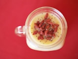 A top-down view of a glass jar filled with a creamy yellow smoothie topped with red goji berries and chia seeds on a vibrant red background.