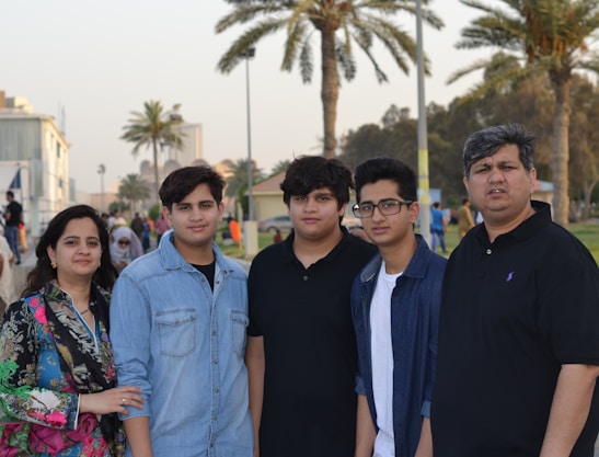 A family stands together outdoors with palm trees and buildings visible in the background. The group consists of two adults and three children, with the mother on the left and the father on the right. They are casually dressed and appear to be in a park or promenade setting.