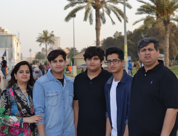 A family stands together outdoors with palm trees and buildings visible in the background. The group consists of two adults and three children, with the mother on the left and the father on the right. They are casually dressed and appear to be in a park or promenade setting.