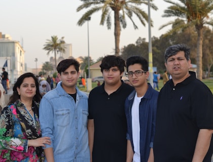 A family stands together outdoors with palm trees and buildings visible in the background. The group consists of two adults and three children, with the mother on the left and the father on the right. They are casually dressed and appear to be in a park or promenade setting.
