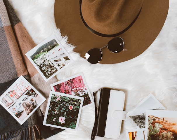 gold-colored framed sunglasses on brown hat beside white and black leather wristlet surrounded by photos on white and gray floral textile