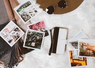 gold-colored framed sunglasses on brown hat beside white and black leather wristlet surrounded by photos on white and gray floral textile