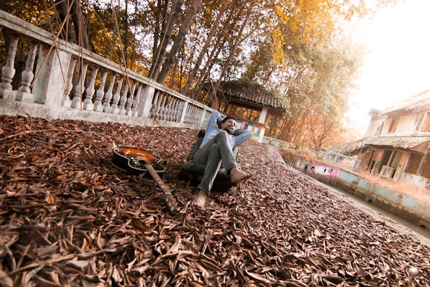 A person is relaxing in a serene outdoor setting, surrounded by fallen leaves, with a guitar lying nearby. The setting includes a stone railing and old buildings, with leaves creating an autumnal atmosphere. Sunlight filters through the trees, casting a warm glow.