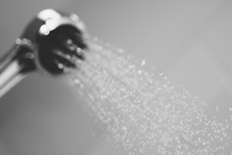 Water sprays out from a metallic showerhead, creating a pattern of droplets against a soft-focus background.