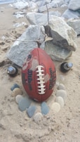 A football is resting upright on sandy beach terrain, encircled by smooth stones. Two beer bottles are positioned on either side, and large rocks are in the background. A twig is tethered to the top of the football.
