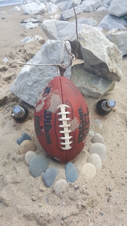 A football is resting upright on sandy beach terrain, encircled by smooth stones. Two beer bottles are positioned on either side, and large rocks are in the background. A twig is tethered to the top of the football.