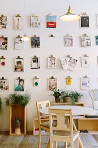 A cozy interior space features a wall adorned with various magazines and pictures clipped onto the wall. There are two hanging lights illuminating the area. Below the wall, a round wooden table with wooden chairs is placed, accompanied by several potted plants and wooden box shelves. The decor creates a relaxed and creative atmosphere.