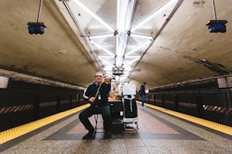 A musician is playing a violin in an underground subway station. The setting features a dimly lit tunnel with fluorescent lights arranged in a geometric pattern on the ceiling. The tiles on the floor are a mix of brown, red, and yellow, with safety strips along the edges. A sign asking for help is visible on the musician's stand, and another person stands in the background looking at a phone.