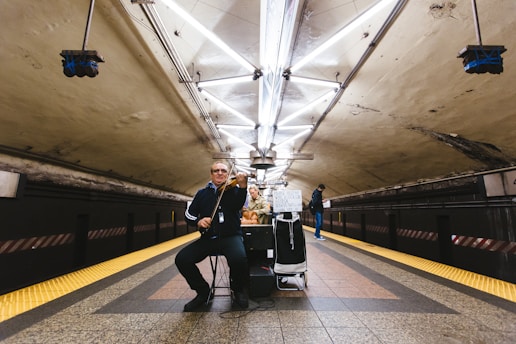 A musician is playing a violin in an underground subway station. The setting features a dimly lit tunnel with fluorescent lights arranged in a geometric pattern on the ceiling. The tiles on the floor are a mix of brown, red, and yellow, with safety strips along the edges. A sign asking for help is visible on the musician's stand, and another person stands in the background looking at a phone.