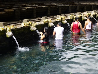 People are participating in a ritual bathing at a water temple, standing in a line under stone spouts with flowing water, surrounded by stone steps and offerings placed on the ledge.