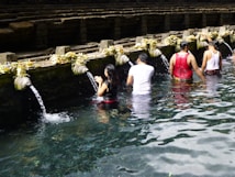 People are participating in a ritual bathing at a water temple, standing in a line under stone spouts with flowing water, surrounded by stone steps and offerings placed on the ledge.