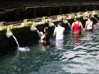 People are participating in a ritual bathing at a water temple, standing in a line under stone spouts with flowing water, surrounded by stone steps and offerings placed on the ledge.
