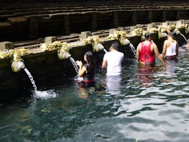 People are participating in a ritual bathing at a water temple, standing in a line under stone spouts with flowing water, surrounded by stone steps and offerings placed on the ledge.