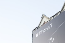 A large billboard prominently displays an advertisement for the iPhone 7 against a clear sky. Part of an ornate building is visible behind the billboard, adding architectural detail to the scene.