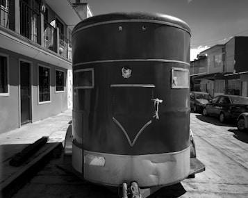 A large, cylindrical trailer is parked on a residential street, with multiple cars lined up along the curb. The buildings in the background have simple, functional architecture with visible balconies and barred windows.