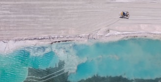 Aerial view of a large turquoise body of water adjacent to a sandy, barren landscape with visible tire tracks, and a construction vehicle near the edge. The scene embodies a contrast between the serene water and the structured, industrialized land.