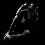 A man sits on the floor, deep in thought with his hand resting on his chin. He wears a plaid shirt and jeans, and his sneakers are visible. The dramatic lighting casts shadows, creating a contemplative and introspective atmosphere.