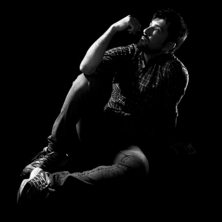 A man sits on the floor, deep in thought with his hand resting on his chin. He wears a plaid shirt and jeans, and his sneakers are visible. The dramatic lighting casts shadows, creating a contemplative and introspective atmosphere.