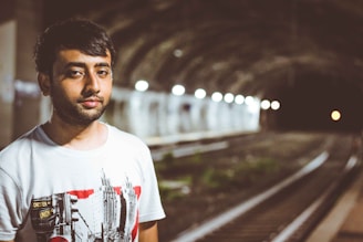 A person stands near the tracks inside a dimly lit underground tunnel. The individual is wearing a light-colored T-shirt with abstract designs and has short dark hair. The tunnel is lined with lights, creating a sense of depth and leading into darkness.