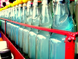 A close-up view of a neatly arranged row of glass bottles filled with a clear liquid, positioned in a metal crate with a red frame. The tops of the bottles are closed, possibly with bottle caps, and there are various colors visible, indicating different flavors or contents. In the background, there are some rounded objects, possibly small fruits, placed atop the bottles.
