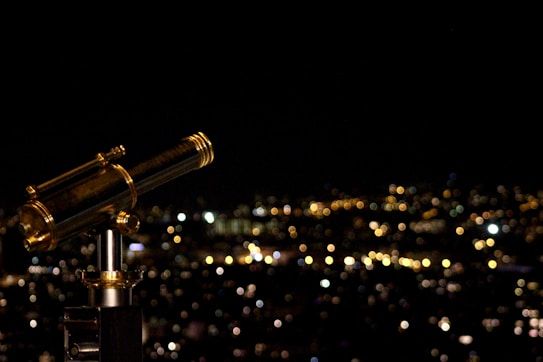 A telescope stands prominently in the foreground with a blurred cityscape in the background, featuring numerous lights creating a bokeh effect against the night sky.