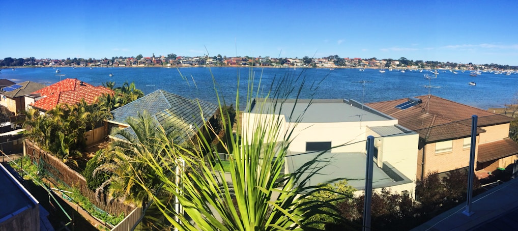 A panoramic view of a waterfront residential area featuring a large body of water with numerous small boats floating in the distance. In the foreground, several modern houses with various colored roofs are visible, surrounded by lush greenery and palm trees. The background displays a long strip of distant urban landscape under a clear blue sky.