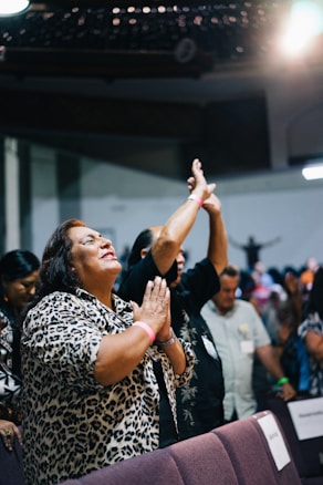 A group of people are gathered indoors, displaying joyful expressions. A woman in the foreground with a leopard print shirt is clasping her hands together, smiling passionately. Other individuals in the background have raised hands, suggesting an engaging and possibly spiritual or celebratory setting.