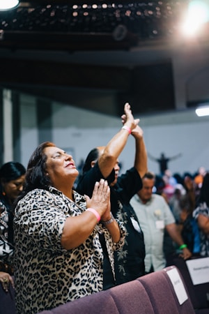 A group of people are gathered indoors, displaying joyful expressions. A woman in the foreground with a leopard print shirt is clasping her hands together, smiling passionately. Other individuals in the background have raised hands, suggesting an engaging and possibly spiritual or celebratory setting.