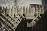 A rustic wooden fence with a lattice pattern features a weathered birdhouse attached to it. A barbed wire is visible atop the fence, and there's a faint outline of a tree shadow on one side.