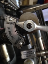 An industrial machine component featuring a metal dial with labels such as 'THIN PAPER' and 'CARDBOARD'. The dial is surrounded by various metal levers and screws, with some text on a small metal plate beneath.