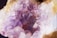 A close-up view of a cluster of amethyst crystals with various shades of purple and white. The crystals have a glossy, translucent appearance with sharp edges and irregular shapes, set against a background of earthy tones.