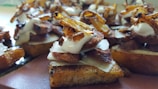 A close-up view of a gourmet appetizer featuring toasted bread topped with slices of roasted meat, caramelized onions, thin cheese slices, and a dollop of creamy sauce. The dish is artistically arranged and appears savory and delicious.