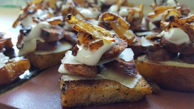 A close-up view of a gourmet appetizer featuring toasted bread topped with slices of roasted meat, caramelized onions, thin cheese slices, and a dollop of creamy sauce. The dish is artistically arranged and appears savory and delicious.
