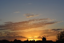 The image captures a serene sunset scene with the sky painted in hues of orange and yellow, transitioning to a darker blue as it reaches the top. Wispy clouds are scattered across the sky, reflecting the warm colors of the setting sun. The silhouette of rooftops and trees line the horizon, adding a sense of calm and peacefulness to the scene.