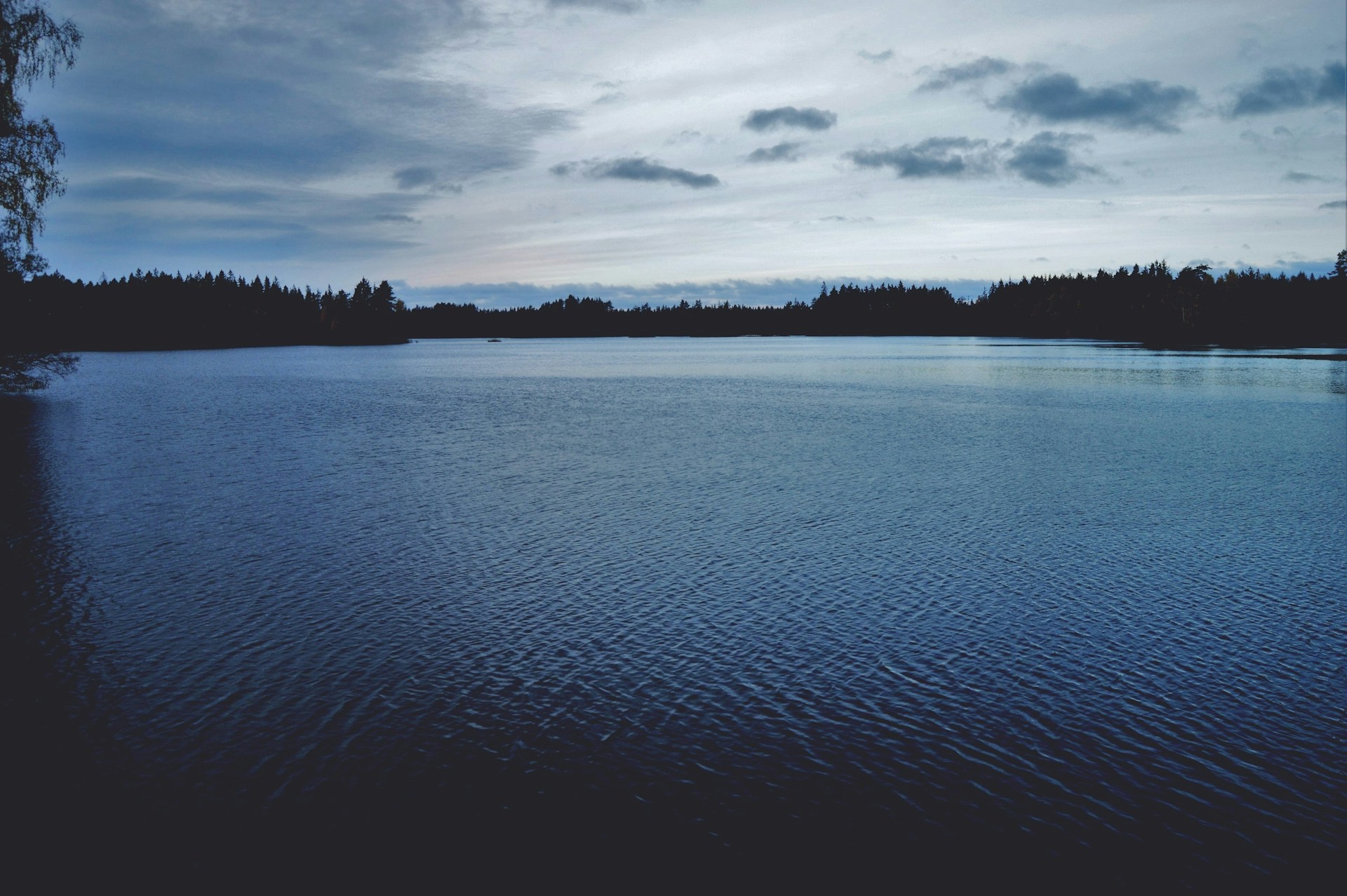 A serene lakeside scene under a cloudy sky, with smooth water reflecting the deep charcoal hues of the surrounding landscape.