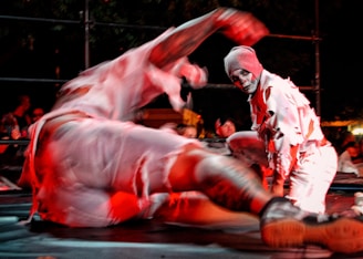 Two performers wearing tattered clothing with one wearing face paint are engaging in a dramatic pose. The scene is illuminated with red lighting, creating a theatrical atmosphere. A crowd in the background watches the performance.