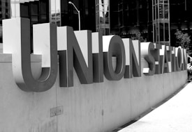 Large, metallic letters spelling 'UNION STATION' are mounted on a concrete wall. The background consists of glass buildings reflecting light, creating a modern urban environment. The image is in black and white, lending a timeless and classic feel.