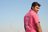 A man wearing a pink sports jersey with the number 7 and the letters FPL on it stands outdoors. The sky is clear, and there are some structures visible in the distant background.
