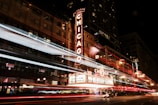 A bustling city street scene at night featuring a prominent theater marquee illuminated with the word 'Chicago'. Light trails from passing vehicles create dynamic streaks across the frame, adding movement to the urban environment. The building's facade features ornate architectural details, and the ambient city lights contribute to a vibrant atmosphere.