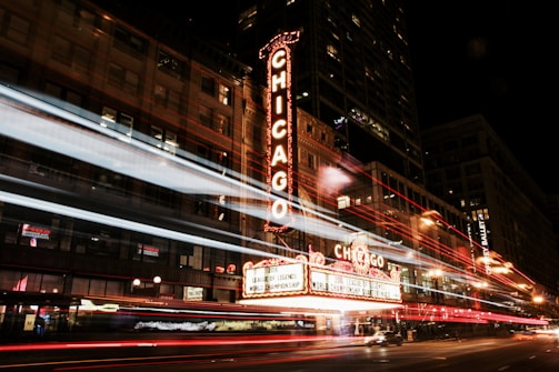 A bustling city street scene at night featuring a prominent theater marquee illuminated with the word 'Chicago'. Light trails from passing vehicles create dynamic streaks across the frame, adding movement to the urban environment. The building's facade features ornate architectural details, and the ambient city lights contribute to a vibrant atmosphere.