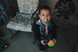A young child with a pacifier clipped to their hoodie is holding colorful toys, standing on a patio near a patterned chair. The child has a pleasant expression, and there is a metal chair nearby with a decorative floral cushion.