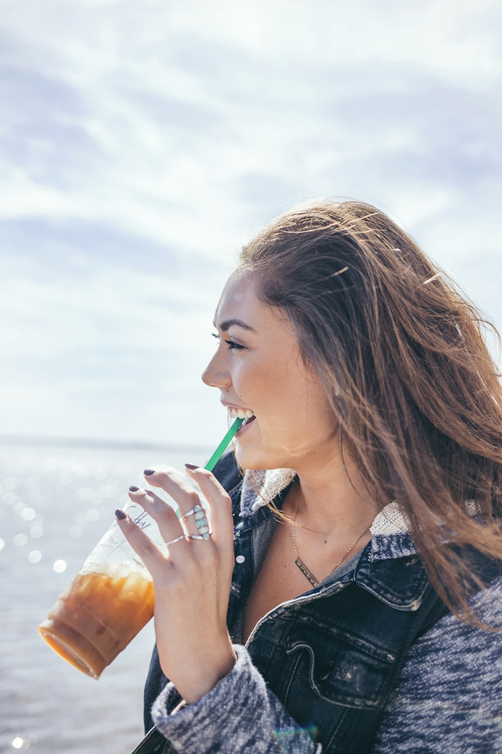 woman wearing black and grey jacket holding glass cup while drinking near body of water during daytime