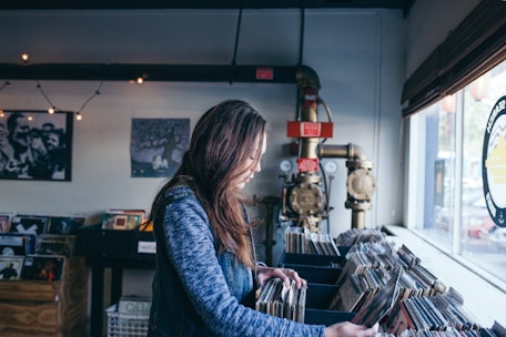 A person with long hair wearing a denim jacket browses through vinyl records in a cozy store. The setting includes various record displays and decorations on the wall, with light coming through the window. Industrial pipes and gauges add to the ambiance.